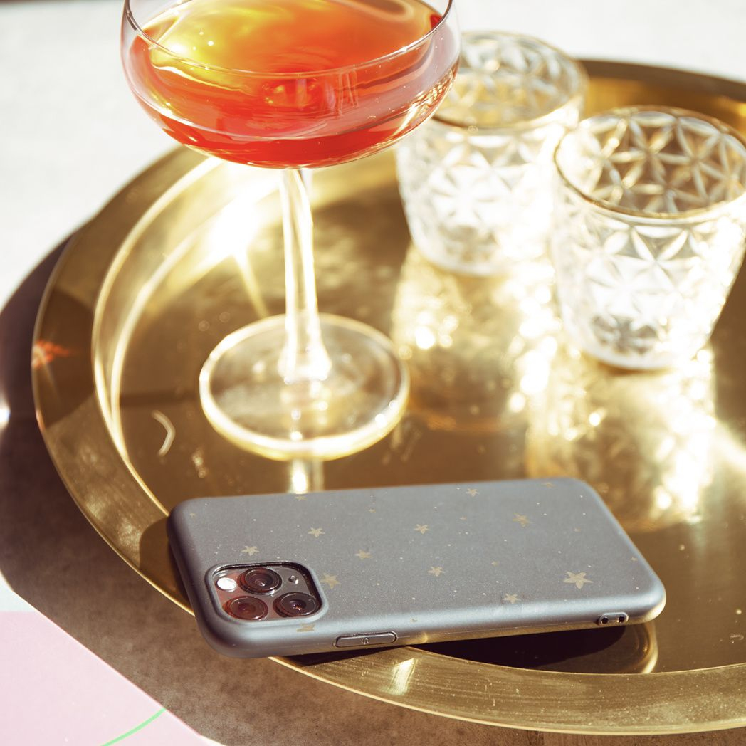 Un cocktail in un bicchiere, due bicchieri decorativi e un telefono con un motivo a stelle su un vassoio dorato. Tutto su una superficie soleggiata.