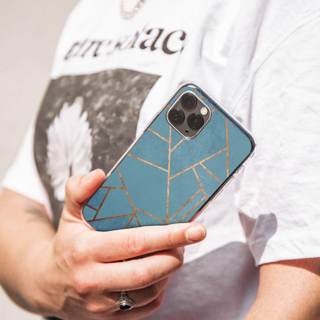 Una persona tiene in mano un telefono con una custodia geometrica blu e oro. Indossa una maglietta bianca con testo e un anello d'argento.