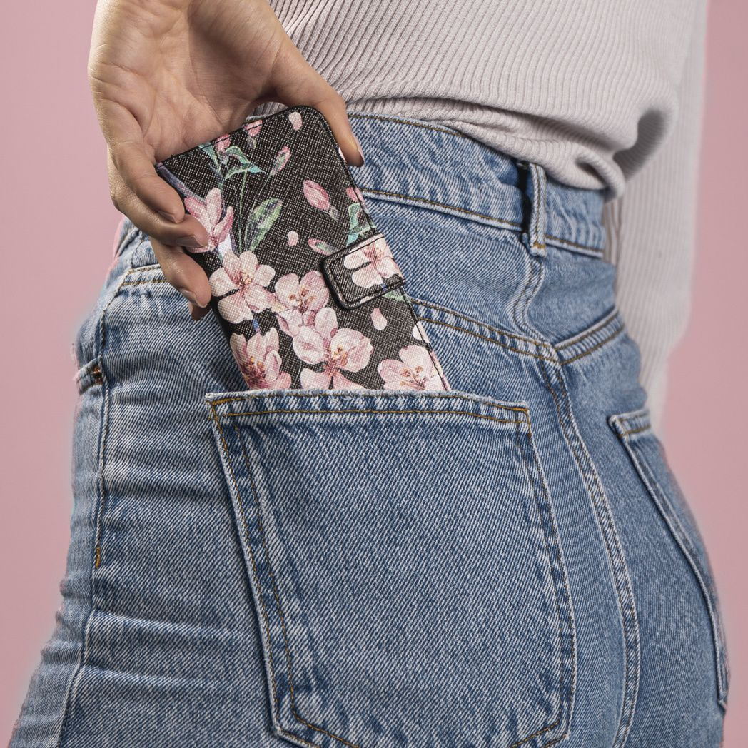 Une main met un étui de téléphone floral dans une poche de jean. L'étui a des fleurs roses. Le jean est bleu clair et le fond est rose.