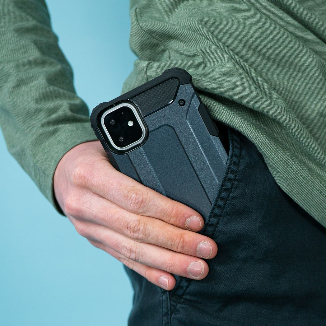De hand van een persoon met een telefoon in een donkergrijze hoes zit in de zak van een donkere broek. De persoon draagt een groen shirt.