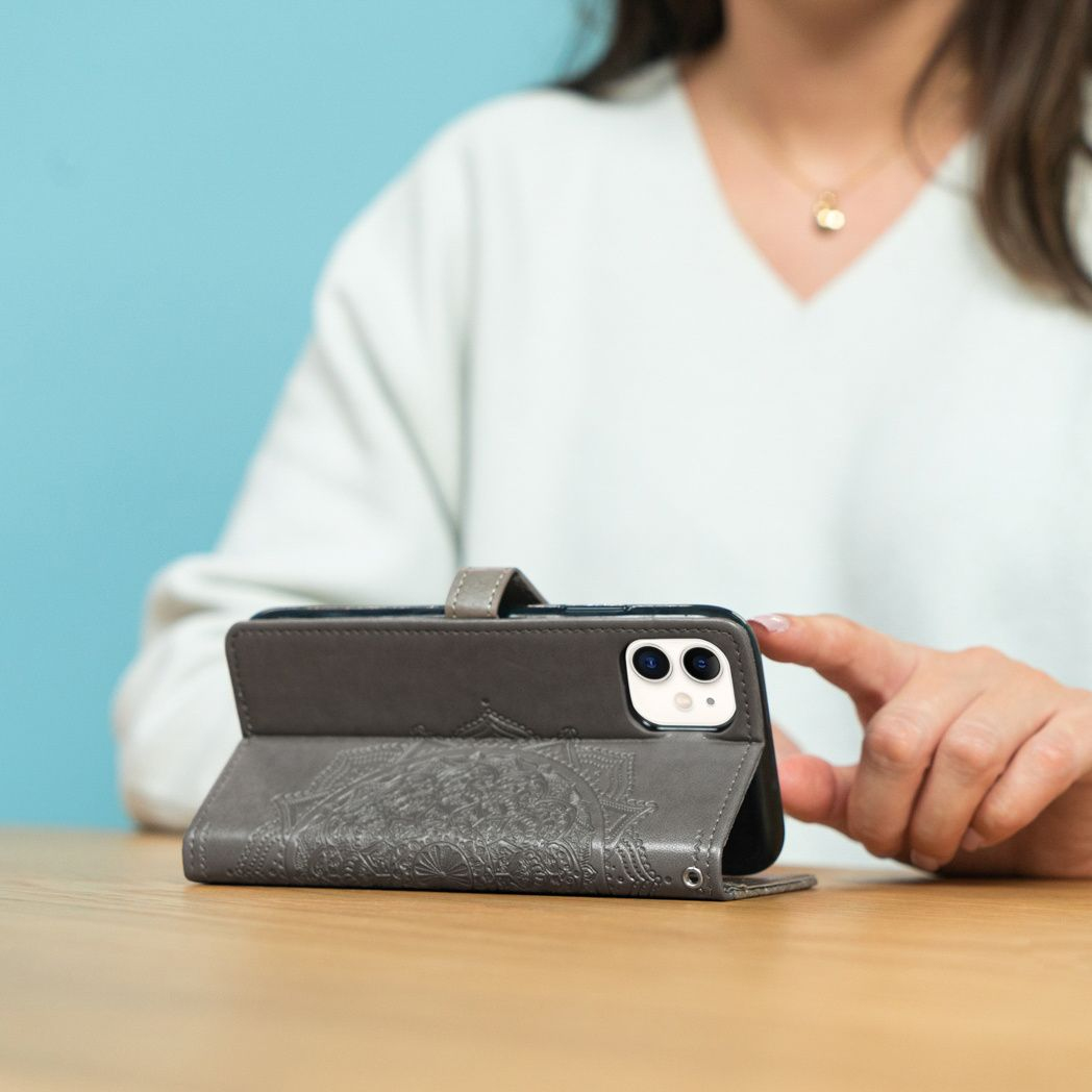 Een vrouw houdt een smartphone vast in een grijze leren hoes met een mandala-ontwerp, op een houten tafel. Een blauwe muur op de achtergrond.