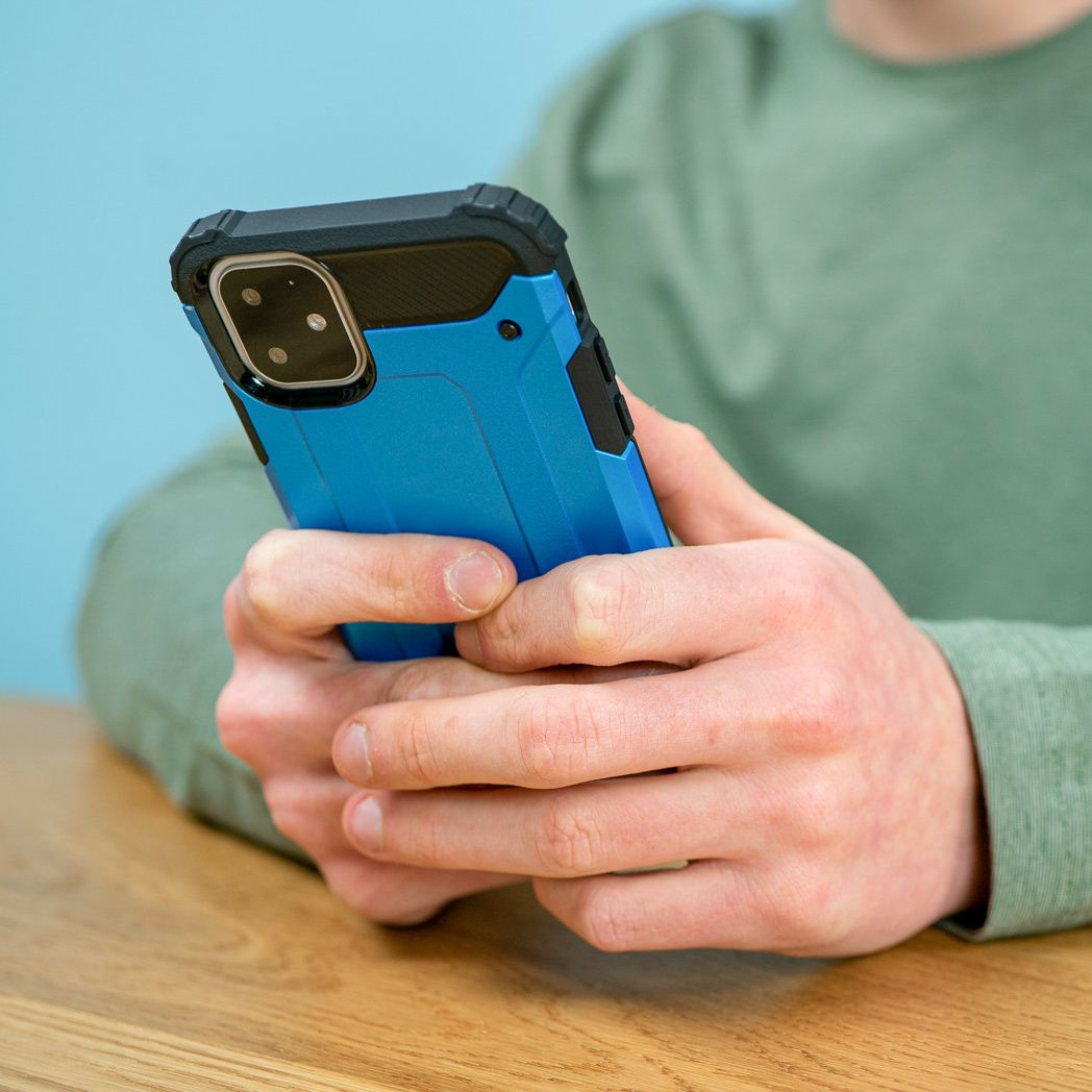 Persoon houdt een blauwe telefoon vast in een beschermhoes. De achtergrond is een lichtblauwe muur en een houten tafel.