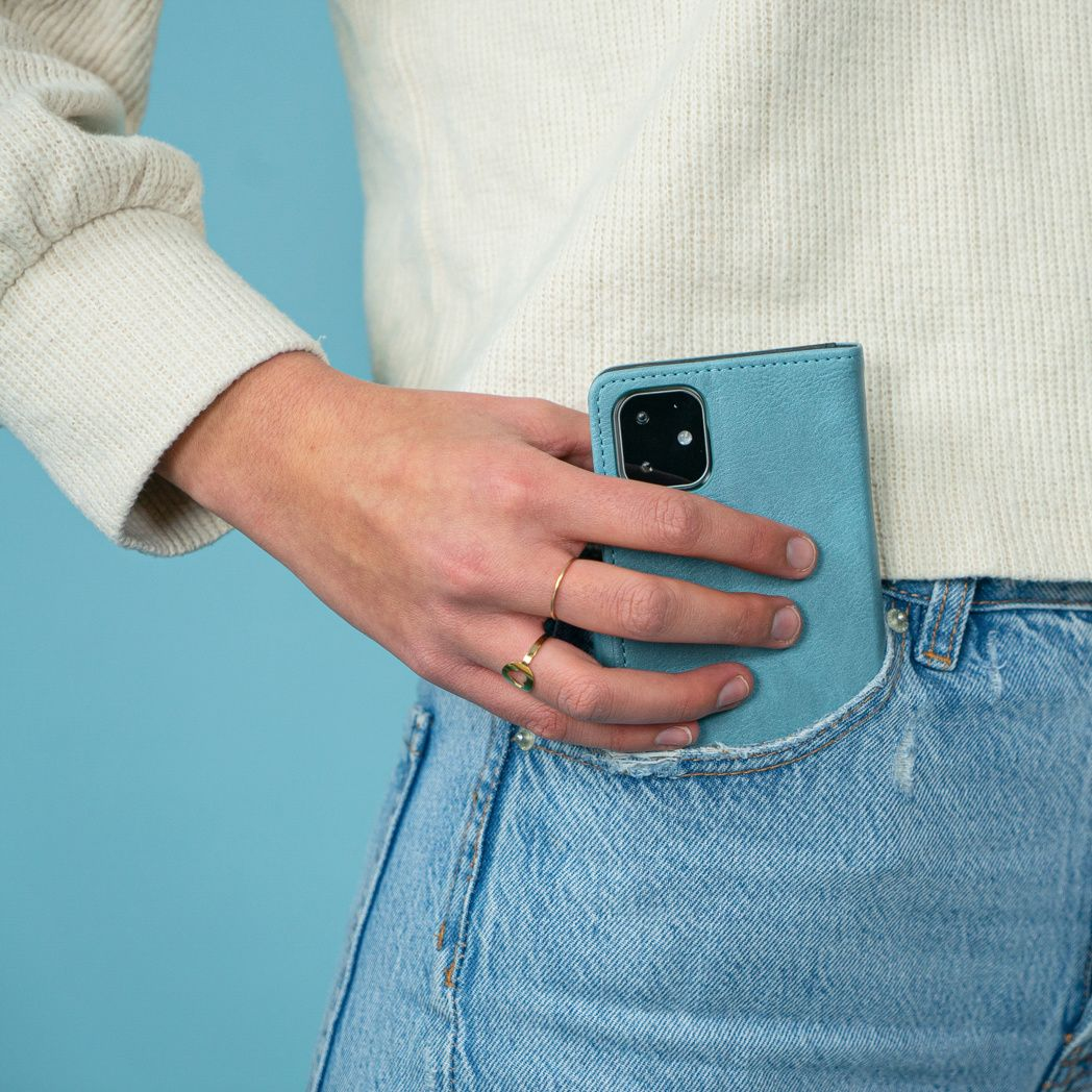 Una persona tiene in tasca una custodia blu per telefono. Indossa un maglione bianco e jeans blu. C'è un anello al dito.
