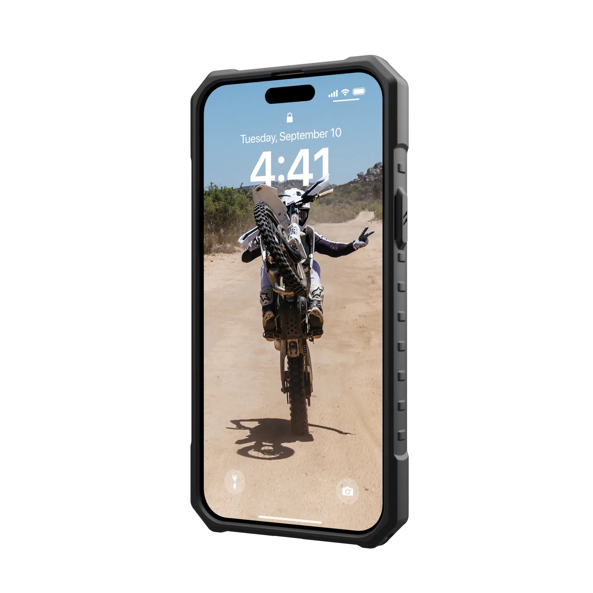Telefon in einer Hülle zeigt einen Dirtbike-Fahrer, der auf einer unbefestigten Straße einen Wheelie macht, sonniger Tag.
