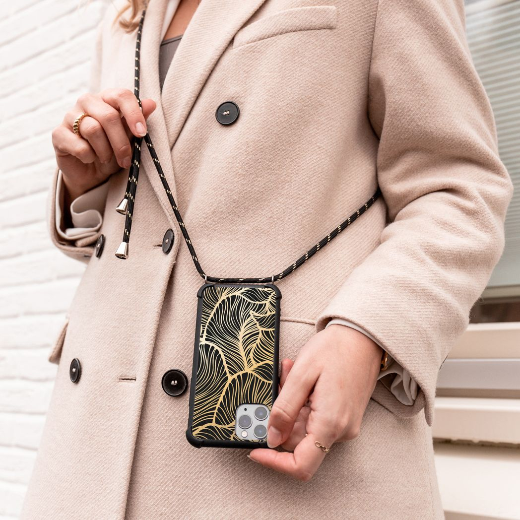 Een vrouw in een beige jas houdt een telefoonhoesje vast met een gouden bladontwerp en een zwarte band. De achtergrond is een witte bakstenen muur.