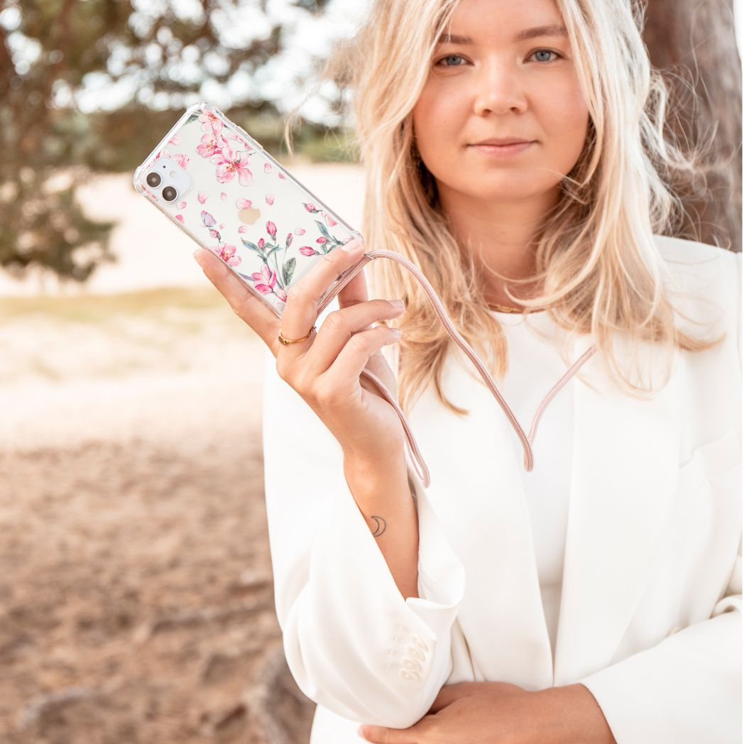Blonde vrouw houdt een telefoon vast in een bloemenhoesje. Ze draagt een witte blazer. Een boom en een veld zijn op de wazige achtergrond.