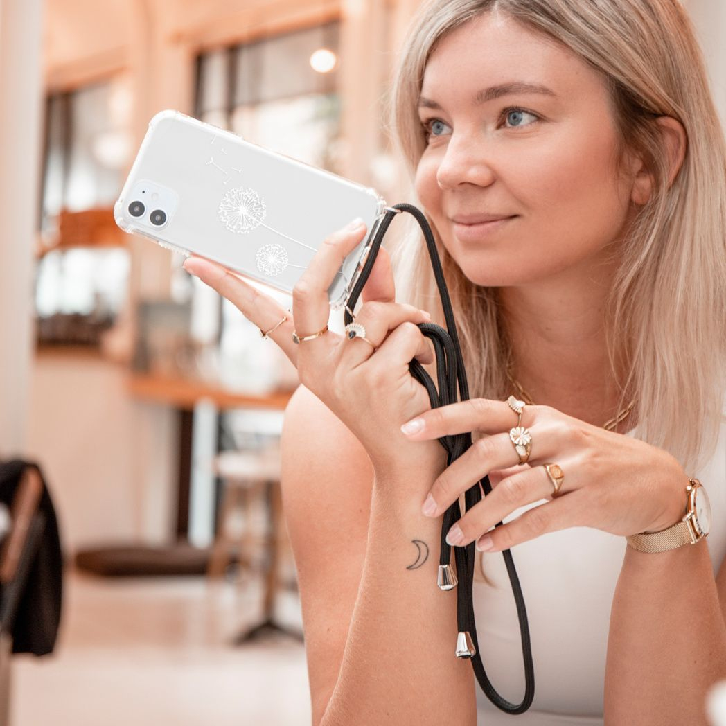 Een vrouw houdt een telefoonhoesje met een paardenbloemontwerp vast. Ze draagt ringen, een horloge en heeft een maantattoo. Blond haar, witte top.