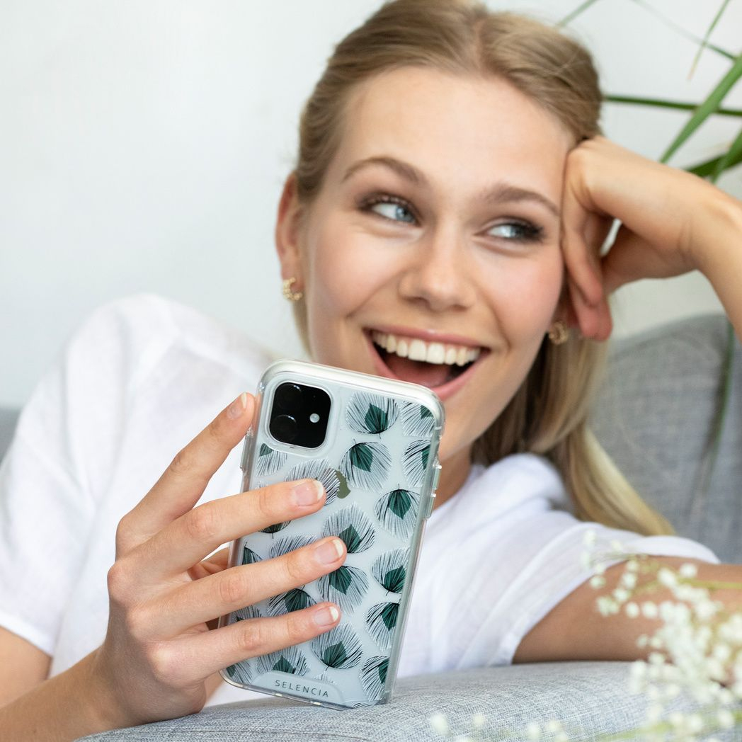 Une femme souriante tient un téléphone avec un étui floral. Elle porte une chemise blanche et des boucles d'oreilles dorées. Elle se repose sur un canapé gris.