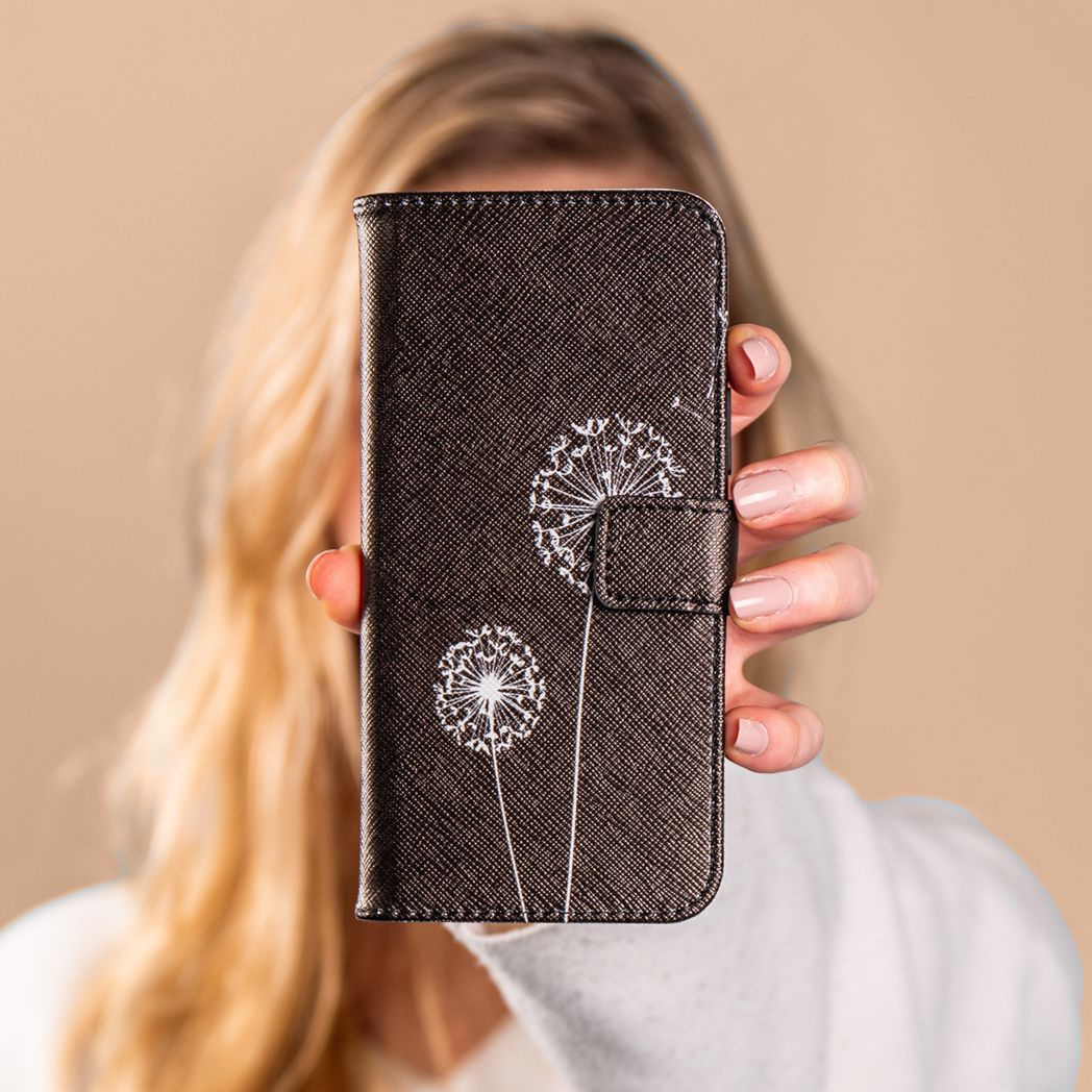 Een vrouw houdt een donker telefoonhoesje vast dat is versierd met witte paardenbloemontwerpen. Haar blonde haar en een witte trui zijn zichtbaar.