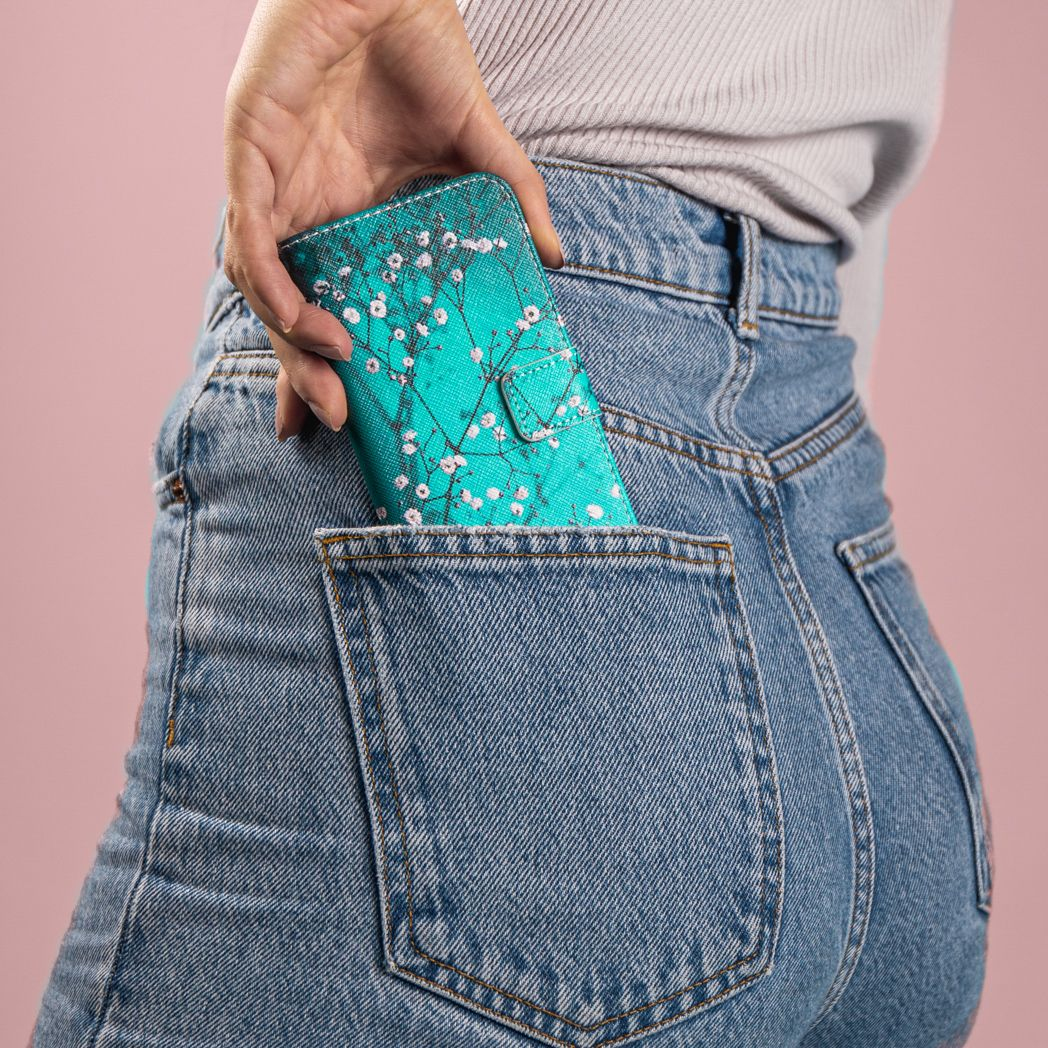 Une main glisse un étui de téléphone vert d'eau avec des fleurs blanches dans une poche de jean. Fond rose et chemise grise.