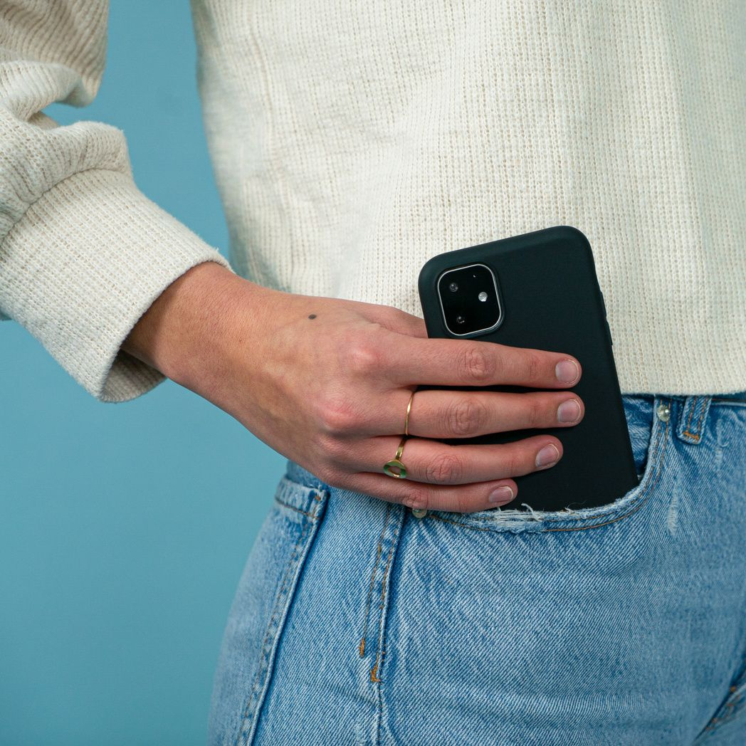 Een persoon met een spijkerbroek en een witte trui houdt een zwarte telefoon in zijn zak. De hand heeft ringen.