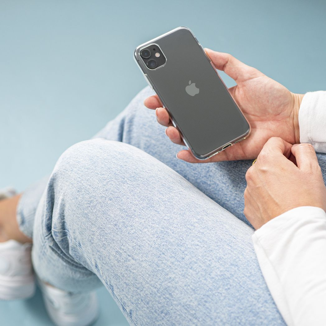 Una persona tiene in mano un iPhone grigio in una custodia trasparente. Indossa jeans e una camicia bianca. Lo sfondo è azzurro.