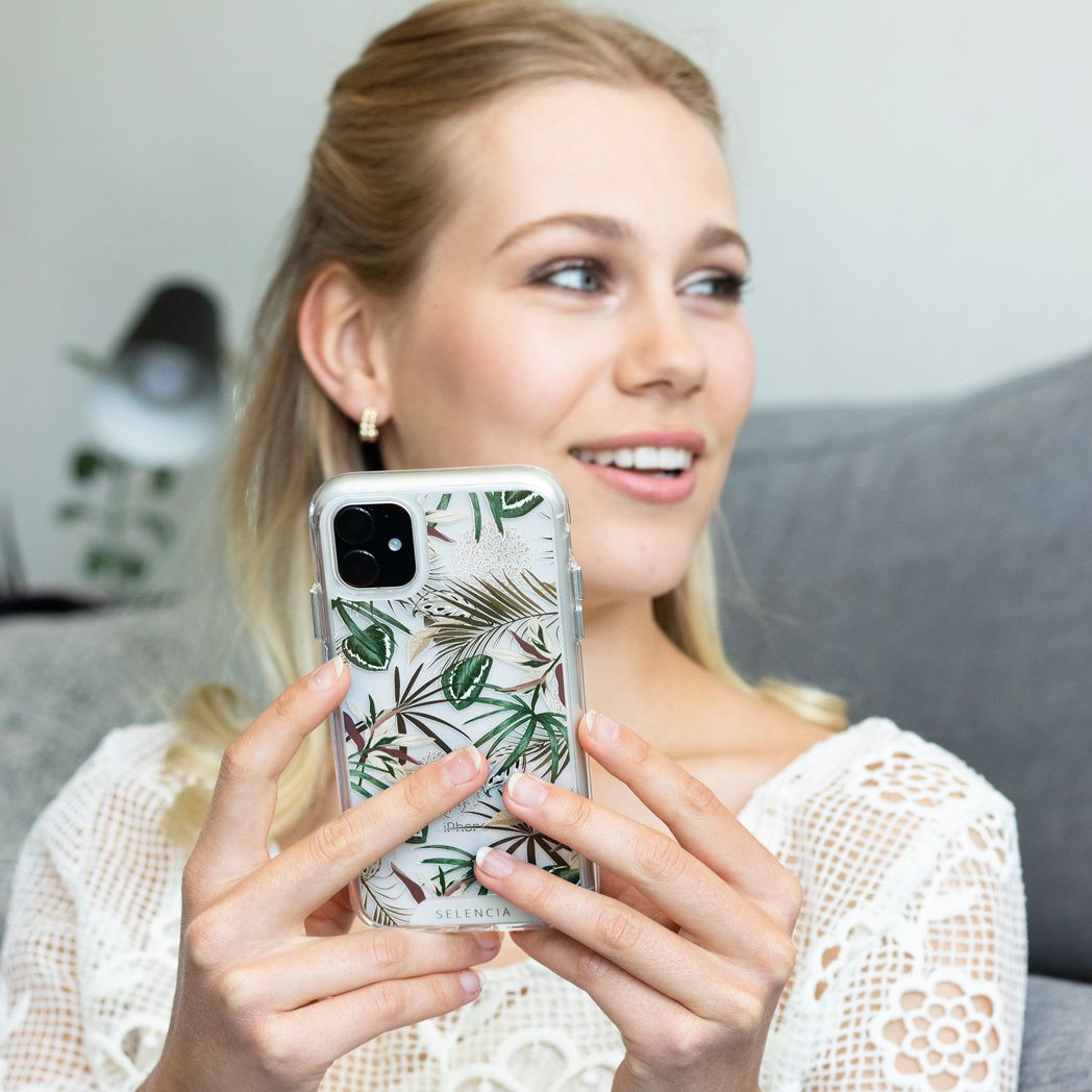 Femme blonde tenant un téléphone avec un étui floral. Elle porte un haut en dentelle blanche et sourit. Le fond est un canapé gris.
