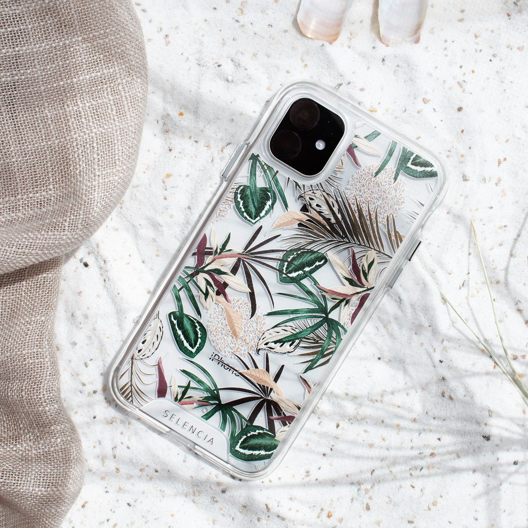 Une coque de téléphone à motifs floraux sur du sable, à côté d'un tissu beige. Coquillages en arrière-plan. La coque dit Selencia.