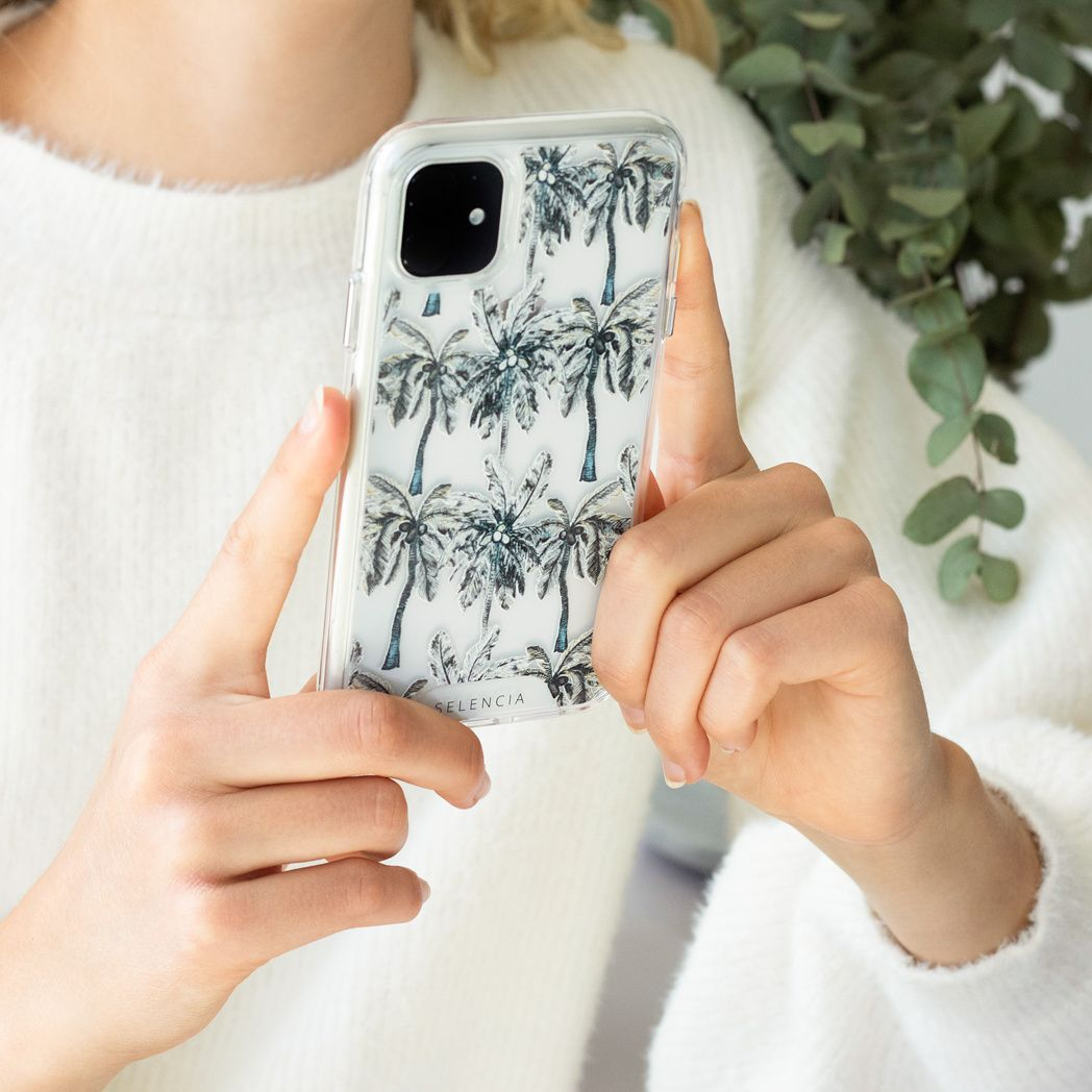 Une personne tient un téléphone avec une coque de palmiers. La personne porte un pull blanc. Des feuilles vertes en arrière-plan.