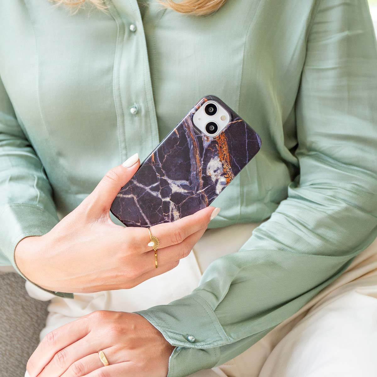 Une femme tient un téléphone avec une coque en marbre. Elle porte une chemise verte et des bagues. L'arrière-plan est flou.