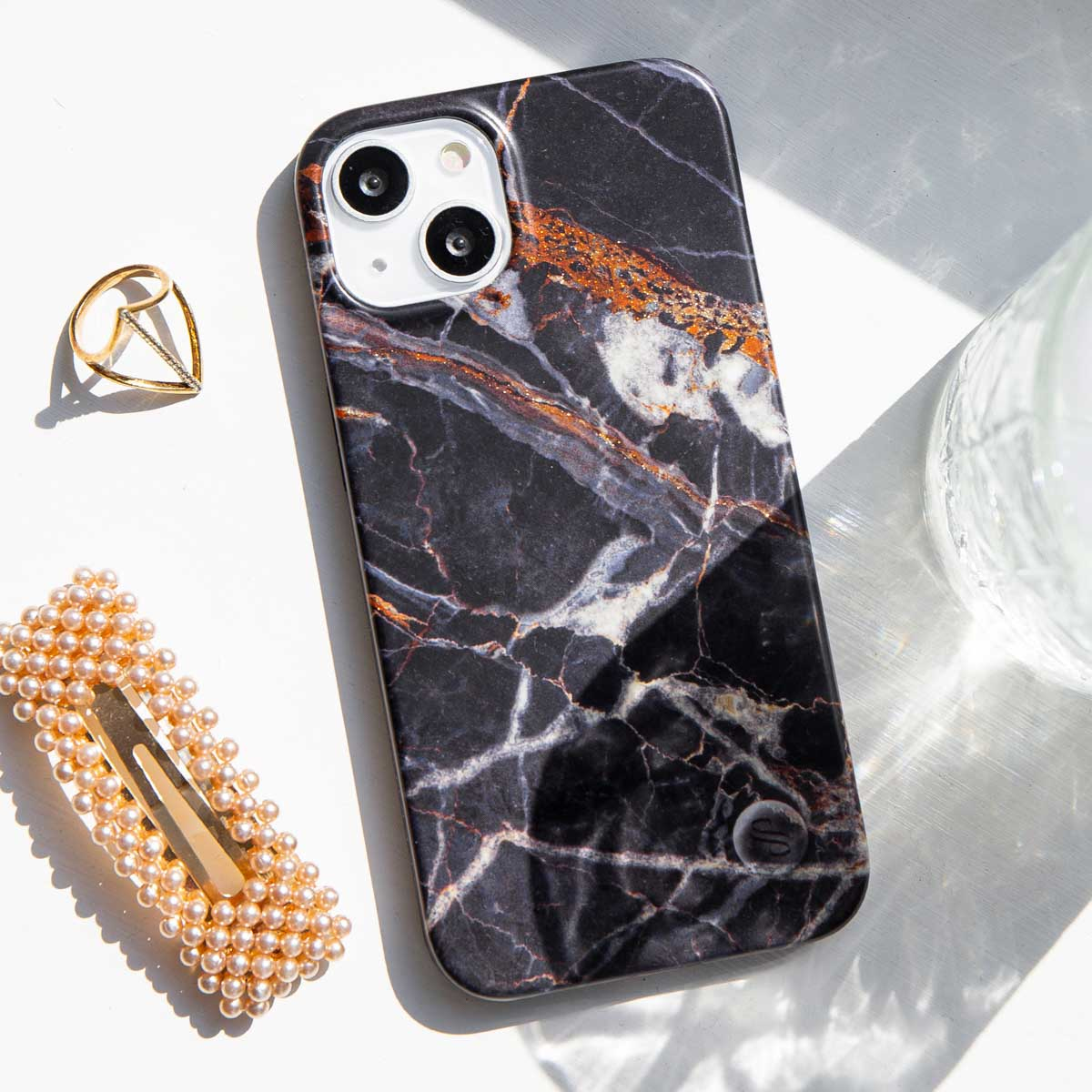 Une coque de téléphone avec un motif marbre, une bague dorée et une pince à cheveux perlée sont sur une surface blanche, près d'un verre.