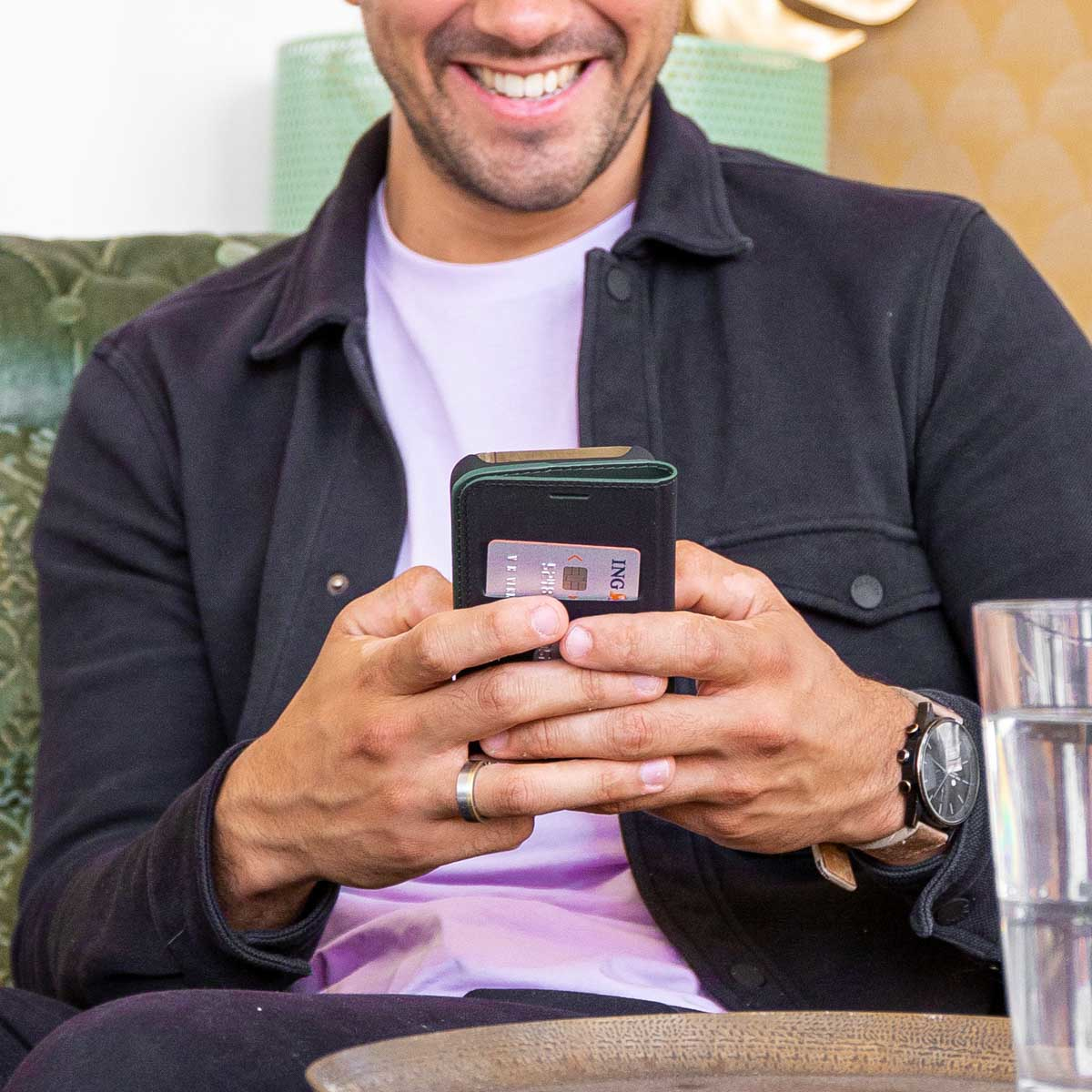 Homme souriant tenant portefeuille et téléphone, montrant une carte bancaire. Il porte une veste noire et une montre. Un verre d'eau à droite.