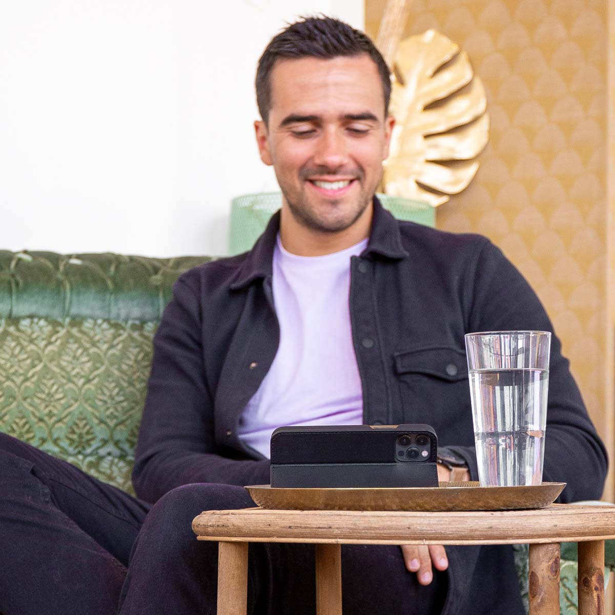 Man lacht, zit op groene bank. Telefoon en glas water op houten tafel voor hem.