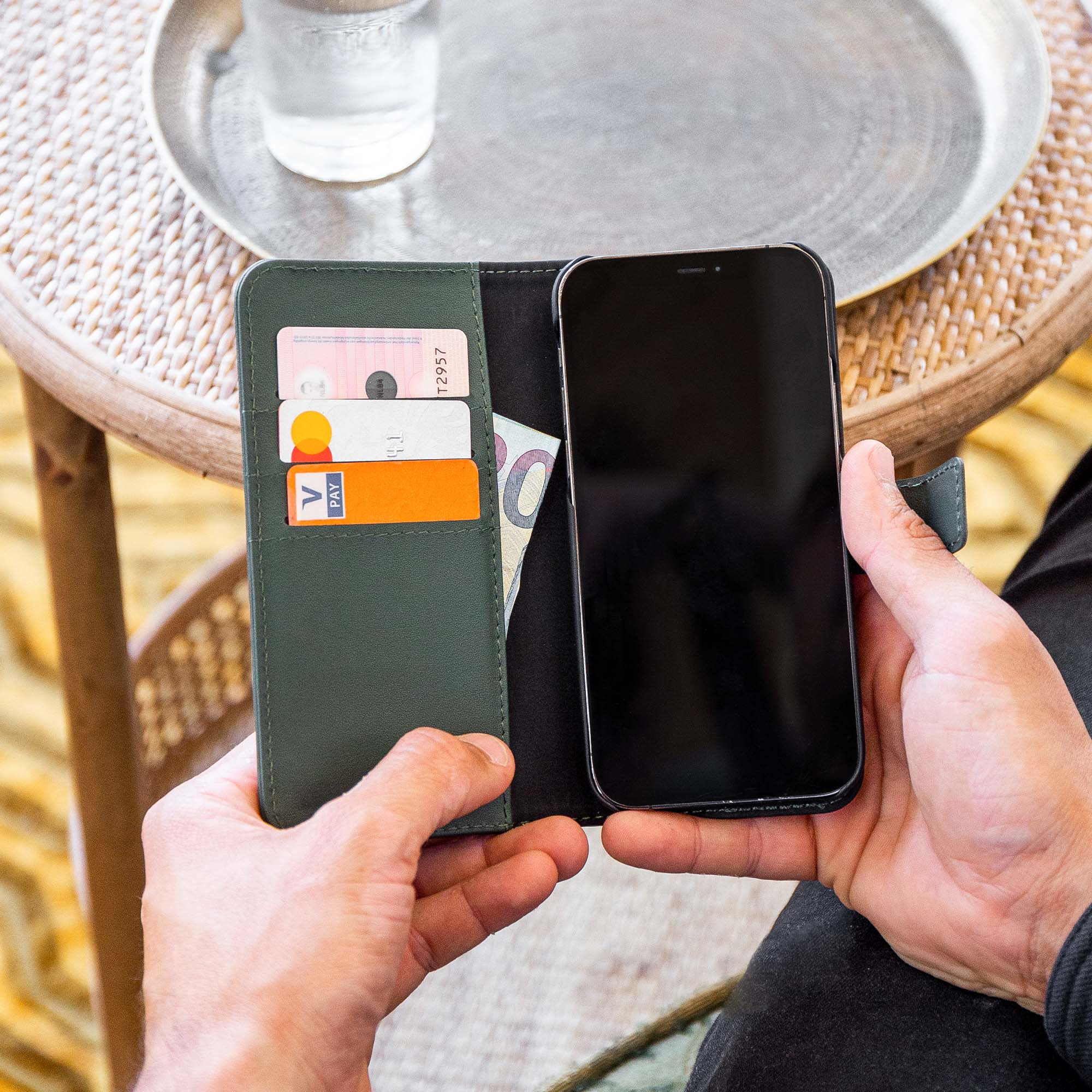 Handen houden een portemonnee met kaarten en een telefoon vast. Een glas water staat op een tafel.