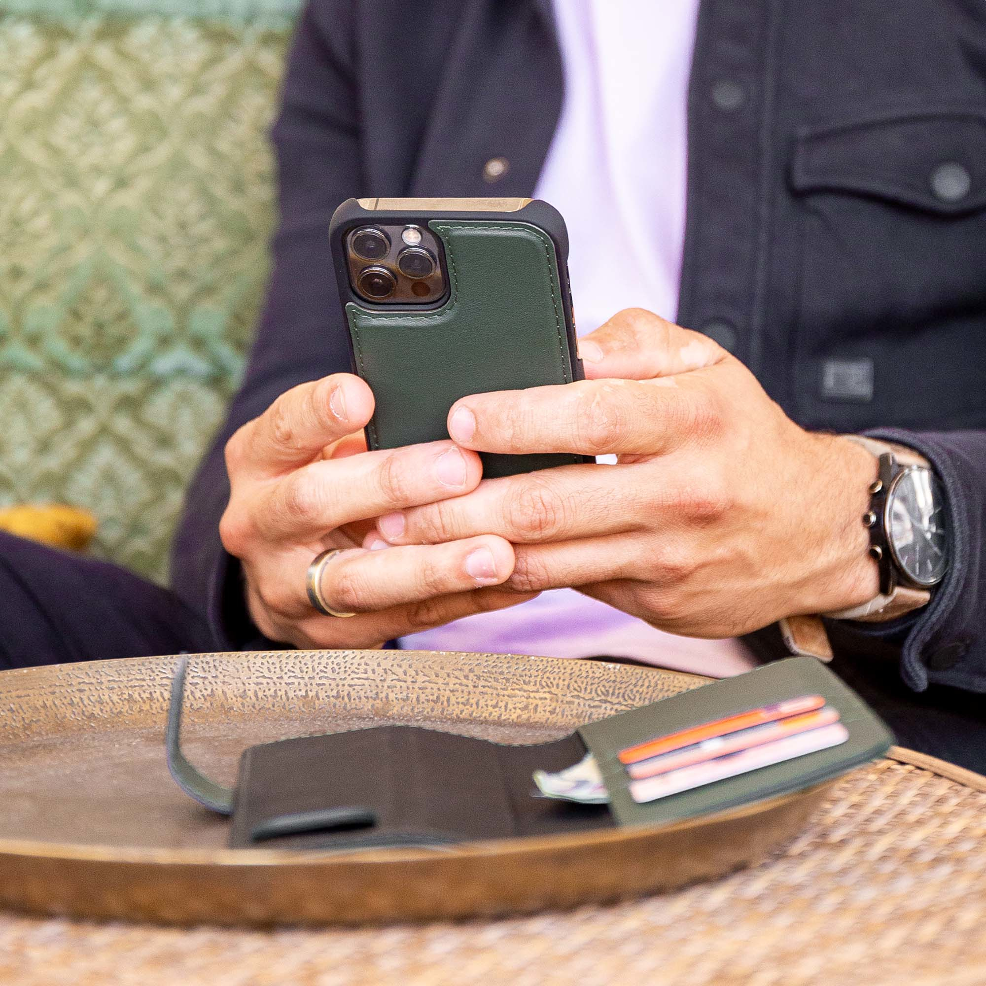Een persoon houdt een telefoon vast in een groene hoes, portemonnee ernaast. De persoon draagt een horloge en ring.