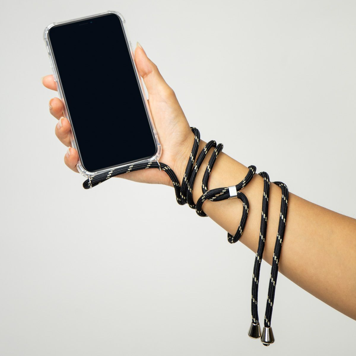 Een hand houdt een telefoon met een zwart scherm in een doorzichtige hoes. De pols is omwikkeld met een zwart-goudkleurig touw.