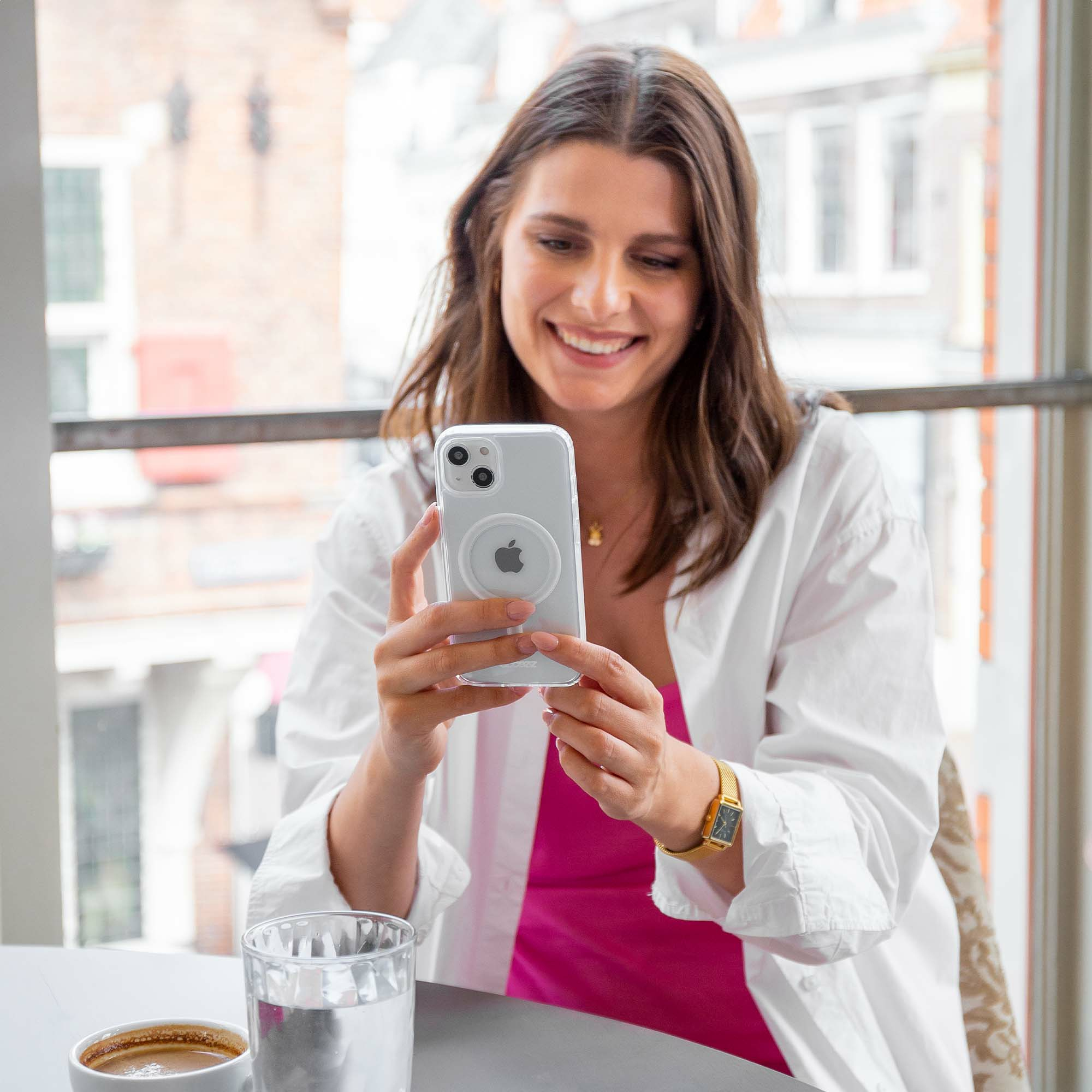 Vrouw glimlacht terwijl ze een telefoon vasthoudt. Ze draagt een wit overhemd, roze top, gouden horloge. Koffiekopje en glas water op tafel.