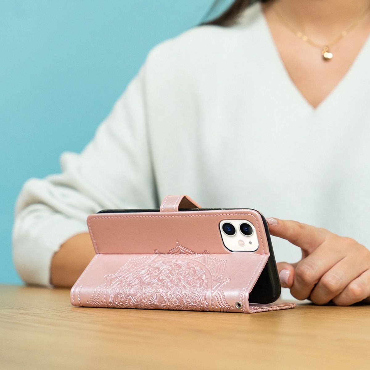 Een vrouw houdt een roze telefoonhoesje vast met een telefoon erin. De hoes zit op een houten tafel. Een blauwe muur is op de achtergrond.
