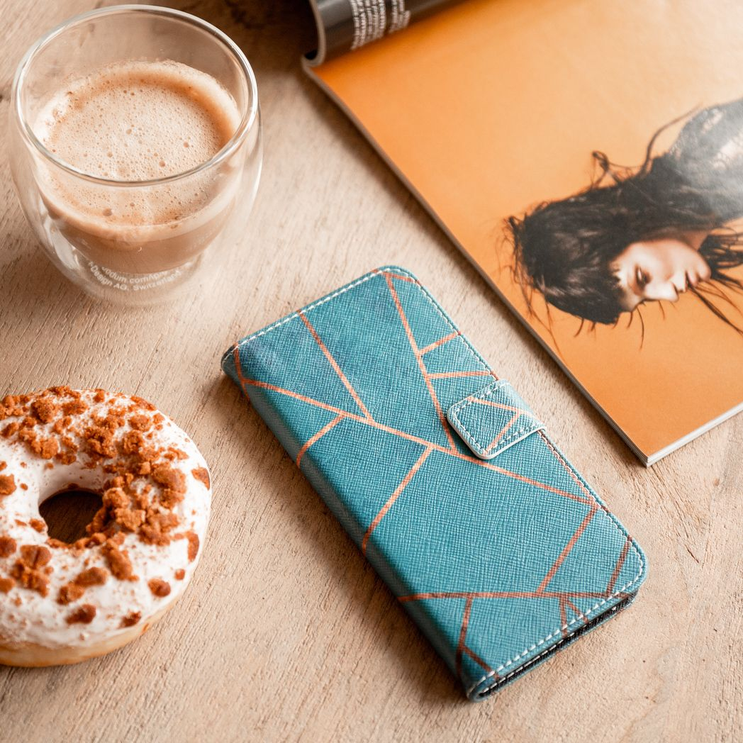 Une table avec un café, un beignet, un étui de téléphone et un magazine ouvert. L'étui est turquoise avec des lignes cuivrées.