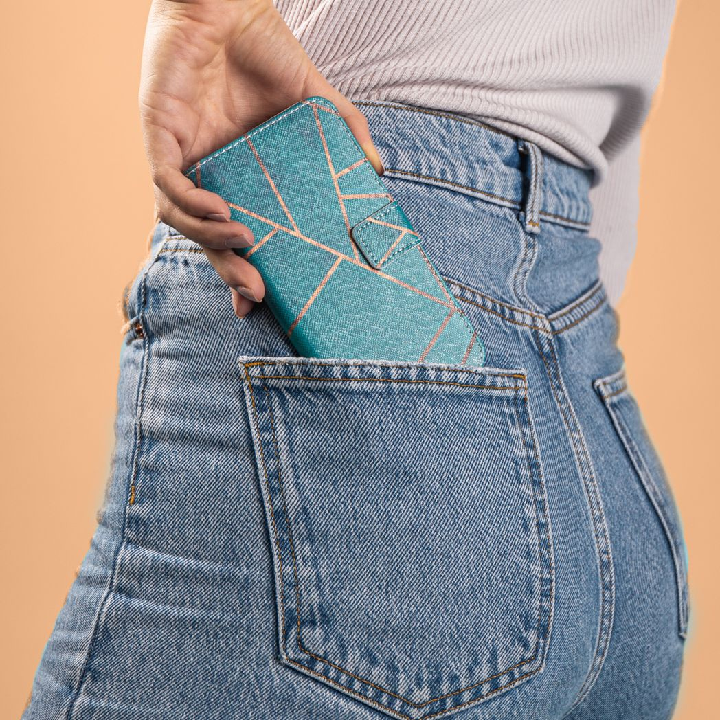 Une personne met un étui de téléphone bleu sarcelle avec un motif géométrique dans une poche arrière de jean. Le fond est pêche.