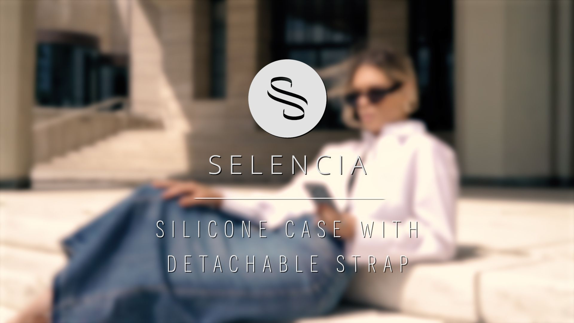 Een vrouw met zonnebril houdt een telefoon vast, zittend buiten. Het SELENCIA-logo is zichtbaar. Tekst: Siliconen hoesje met afneembare band.