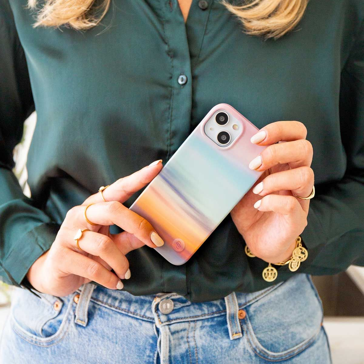 Une personne tient un téléphone avec un étui coloré. Elle porte des bagues, un bracelet, une chemise vert foncé et un jean.