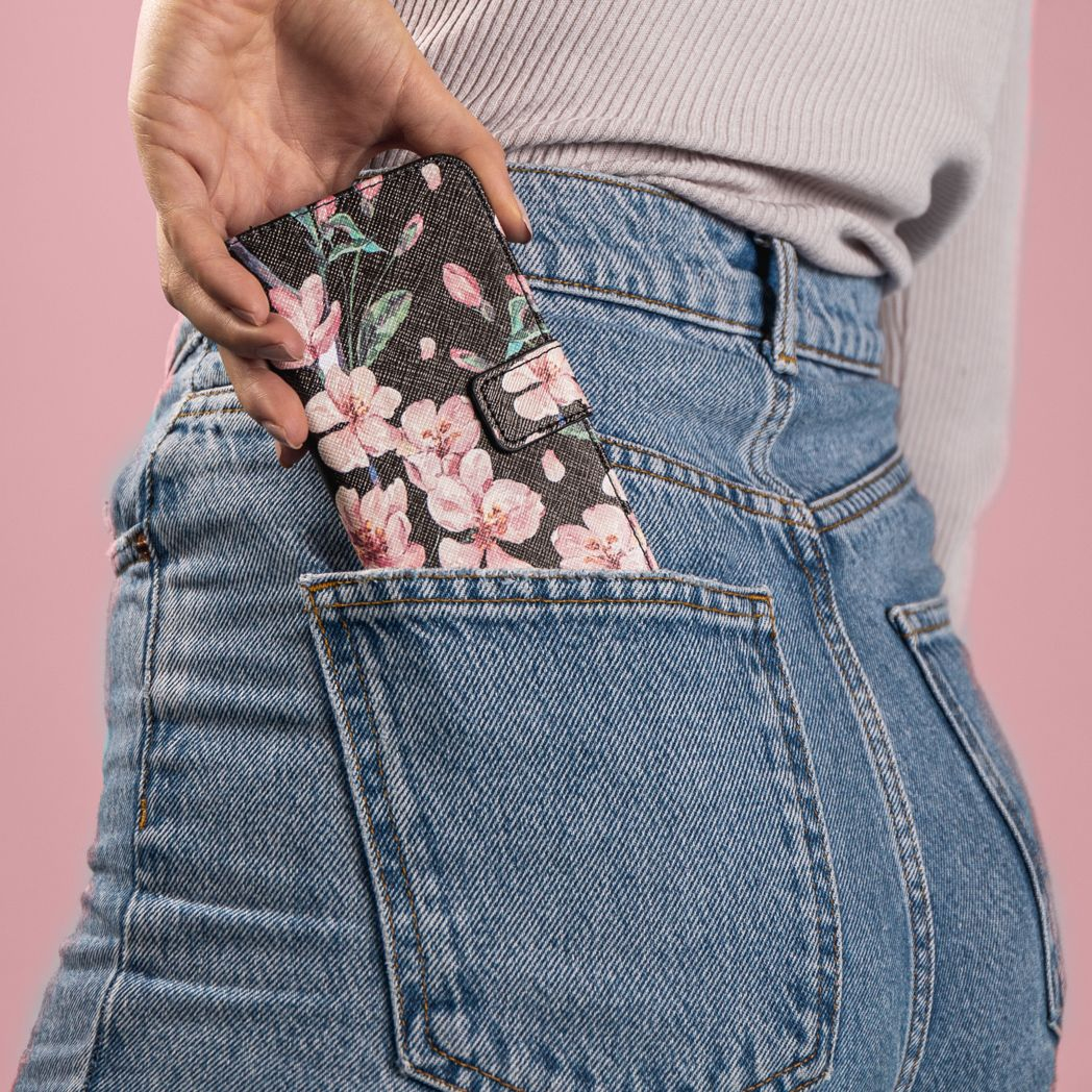 Une main insère un étui de téléphone à motifs floraux dans une poche de jean. L'étui est noir avec des fleurs roses. Le fond est un mur rose.