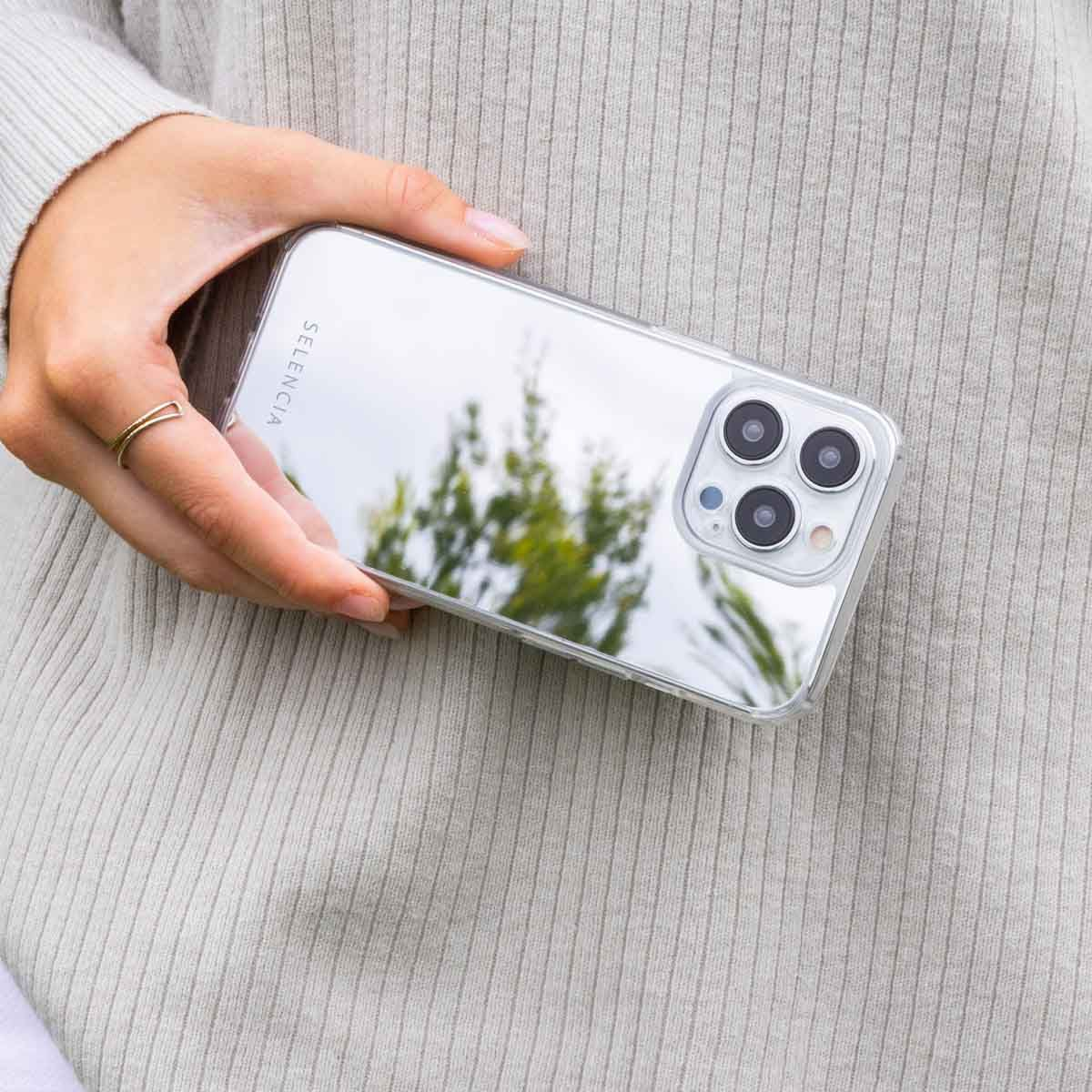 Eine Hand hält eine transparente Handyhülle. Die Hülle spiegelt Grün wider. Die Person trägt einen Ring und einen grauen Pullover.