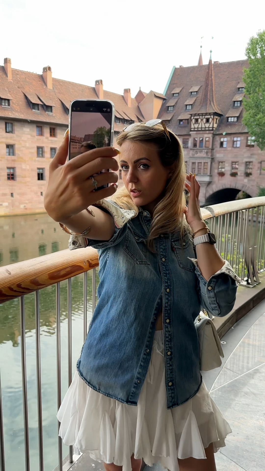Frau macht ein Selfie mit einem Telefon, trägt ein Jeanshemd und einen weißen Rock. Im Hintergrund sind alte Gebäude und ein Fluss zu sehen.