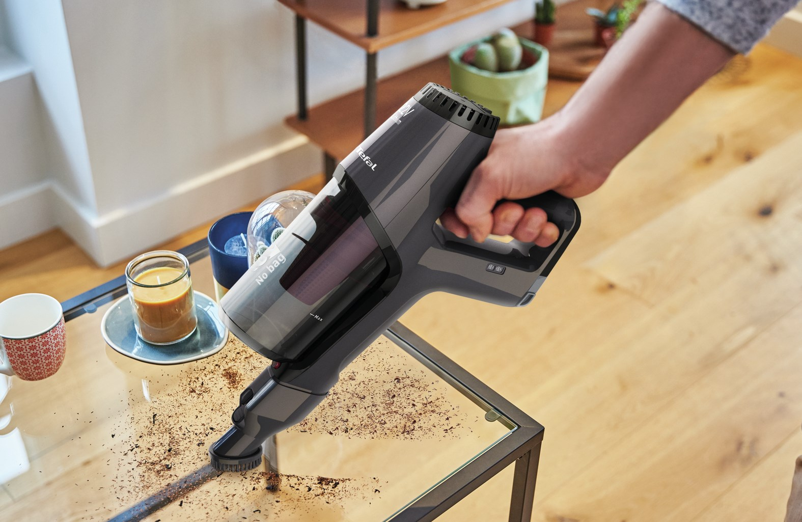 Ręka trzyma szary odkurzacz Tefal nad szklanym stołem z zanieczyszczeniami. Świece i dekoracje w tle.