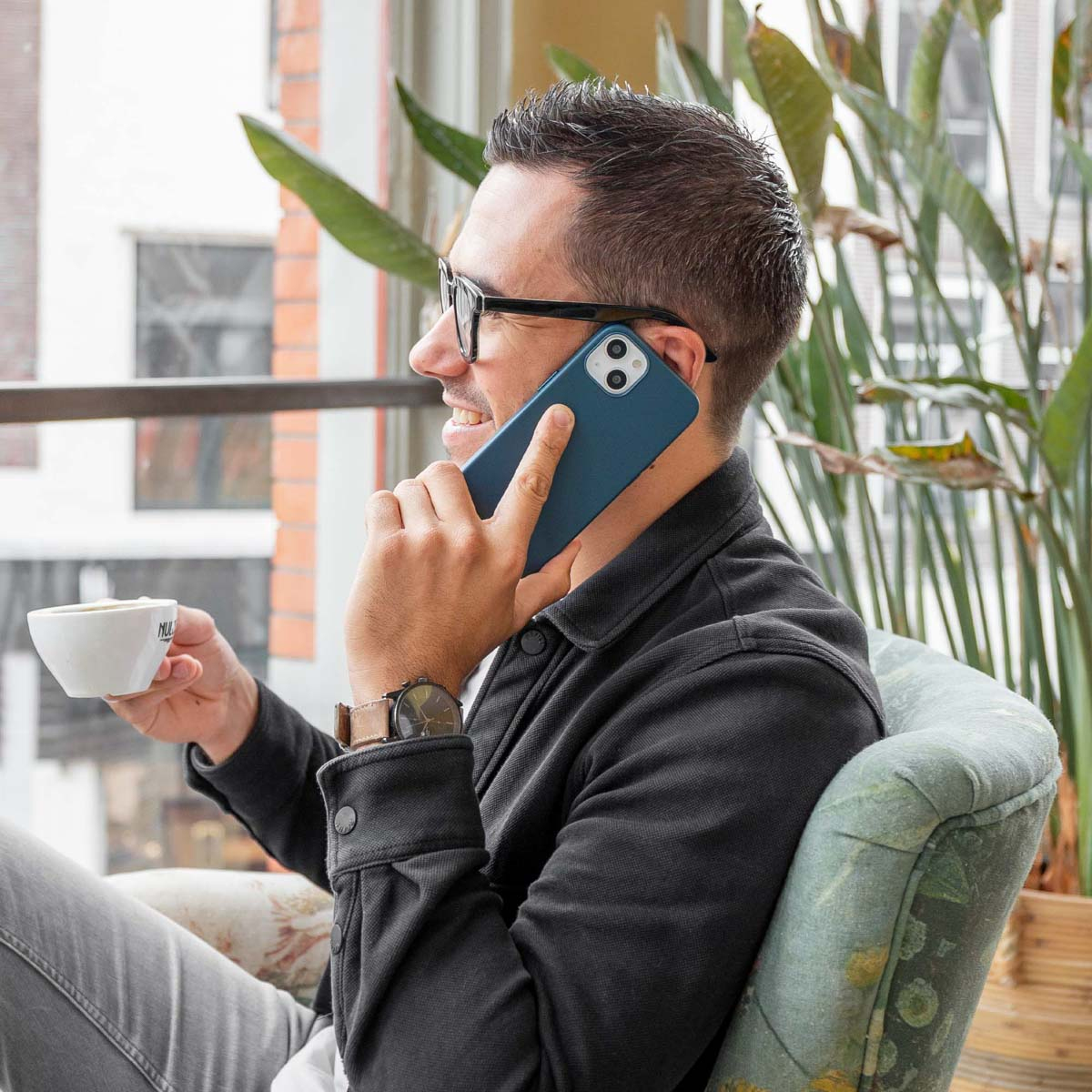 Man met bril telefoneert, houdt koffie vast, zit in een stoel, met planten en een raam op de achtergrond.