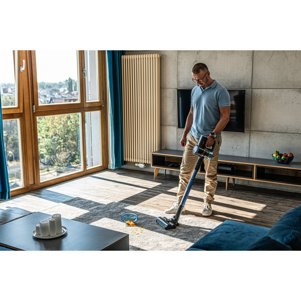 Mężczyzna odkurza dywan w salonie. Jest okno, telewizor i sofa. Rozrzucone jedzenie i miska leżą na dywanie.