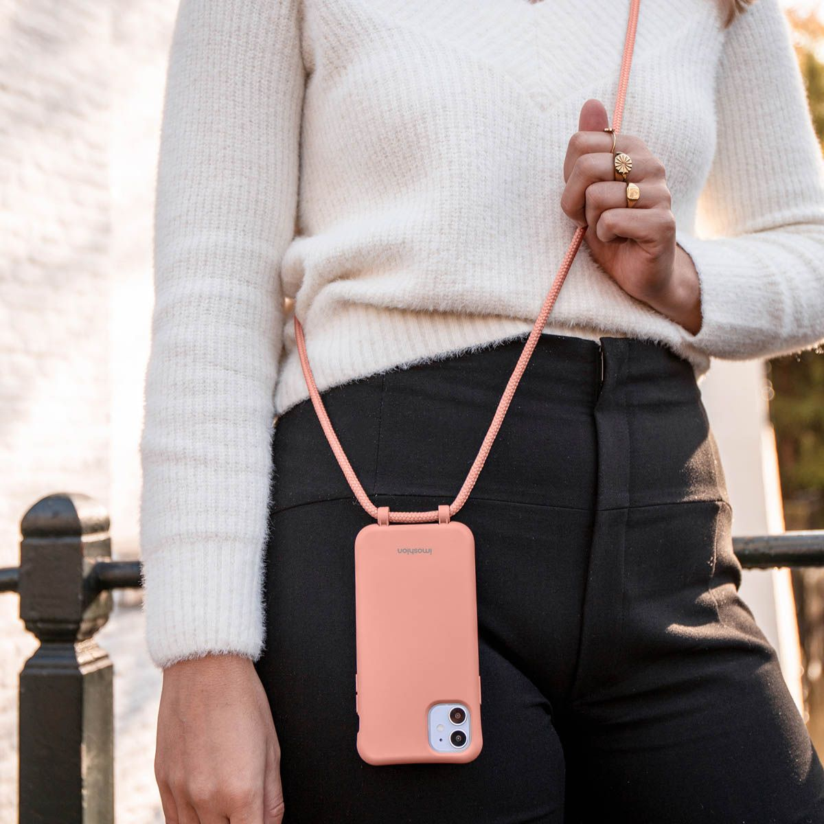 Een vrouw draagt een perzikroze telefoonhoesje met een bijpassende riem. Ze draagt een witte trui en een zwarte broek. Gouden ringen sieren haar vingers.