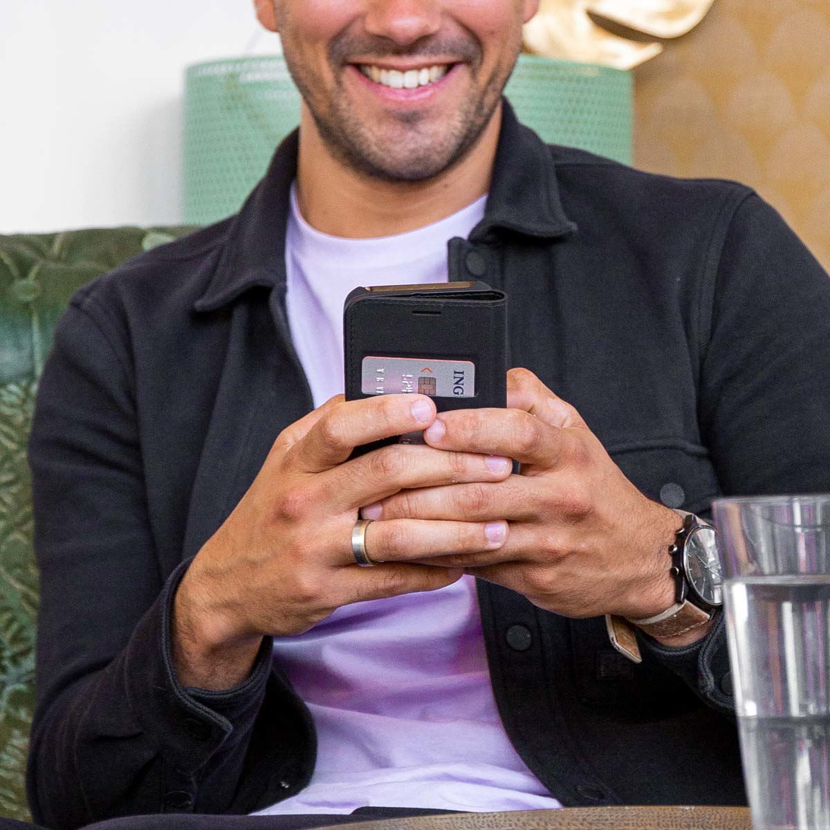 Un homme souriant tient un téléphone avec une carte de crédit. Il porte une veste noire et une montre. Le fond est flou.