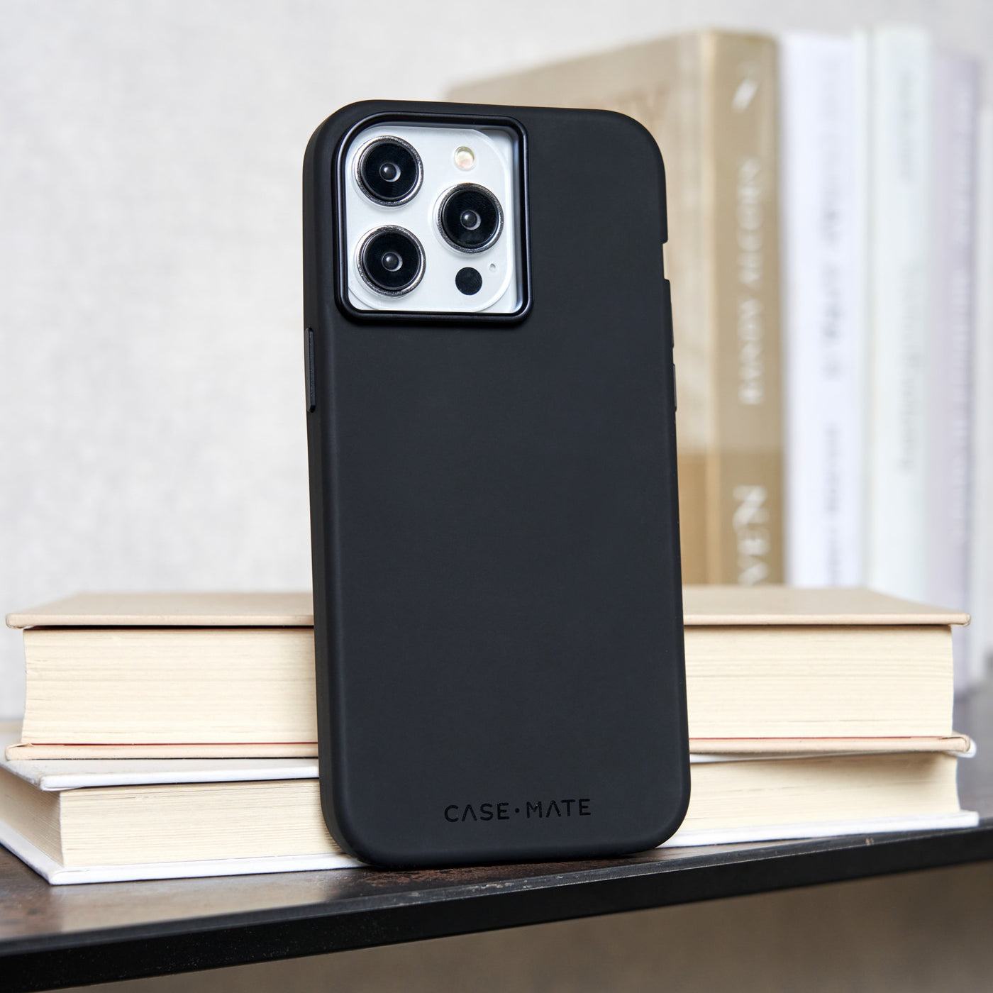 Un iPhone nero con custodia è appoggiato su libri. Il telefono ha una sporgenza per la fotocamera. I libri sono beige e bianchi.
