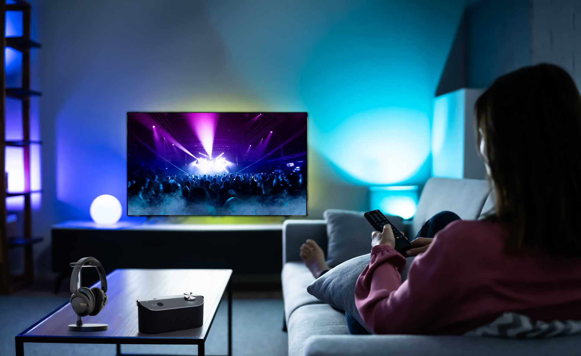 Eine Person sieht sich ein Konzert im Fernsehen an. Kopfhörer und ein Lautsprecher stehen auf einem Tisch. Der Raum hat Umgebungslicht.