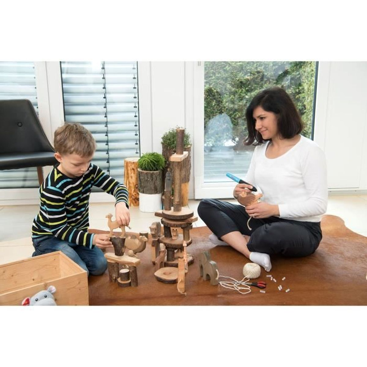 Eine Frau und ein Kind spielen mit Holzspielzeug auf einem braunen Teppich in der Nähe eines Fensters. Das Kind trägt ein gestreiftes Hemd.