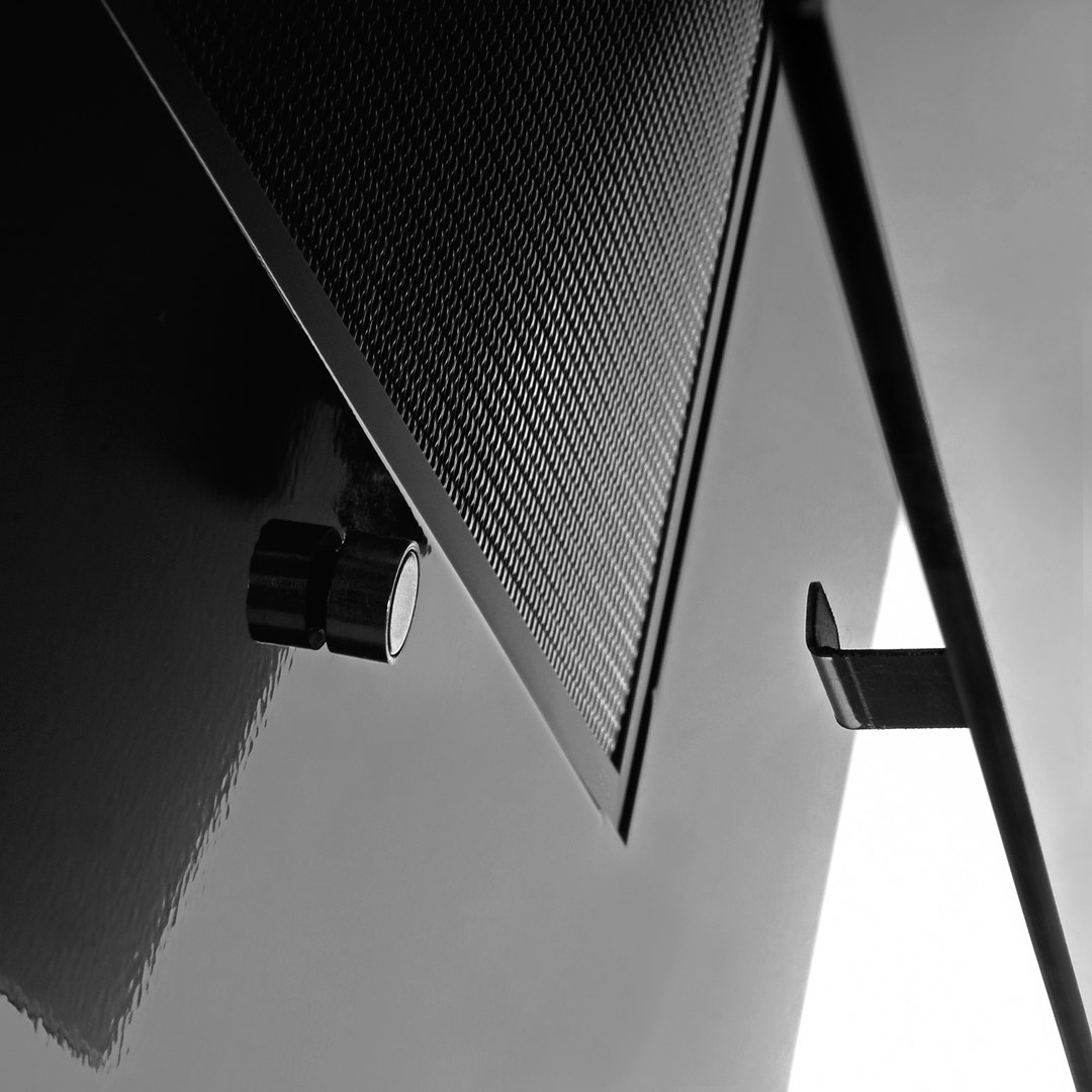 Zbliżenie czarnego obiektu z metalową siatką, dwoma pokrętłami i wspornikiem. Tło jest szare i białe.