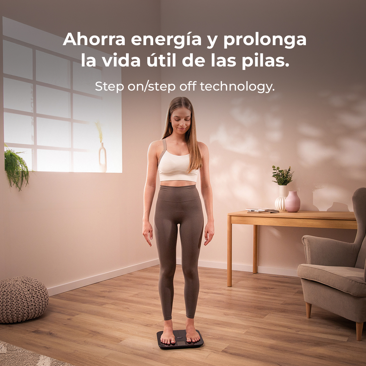 Una mujer está de pie sobre una balanza en interiores. Viste top blanco y leggings grises. El texto superior dice: Ahorra energía y prolonga la vida útil de la batería.