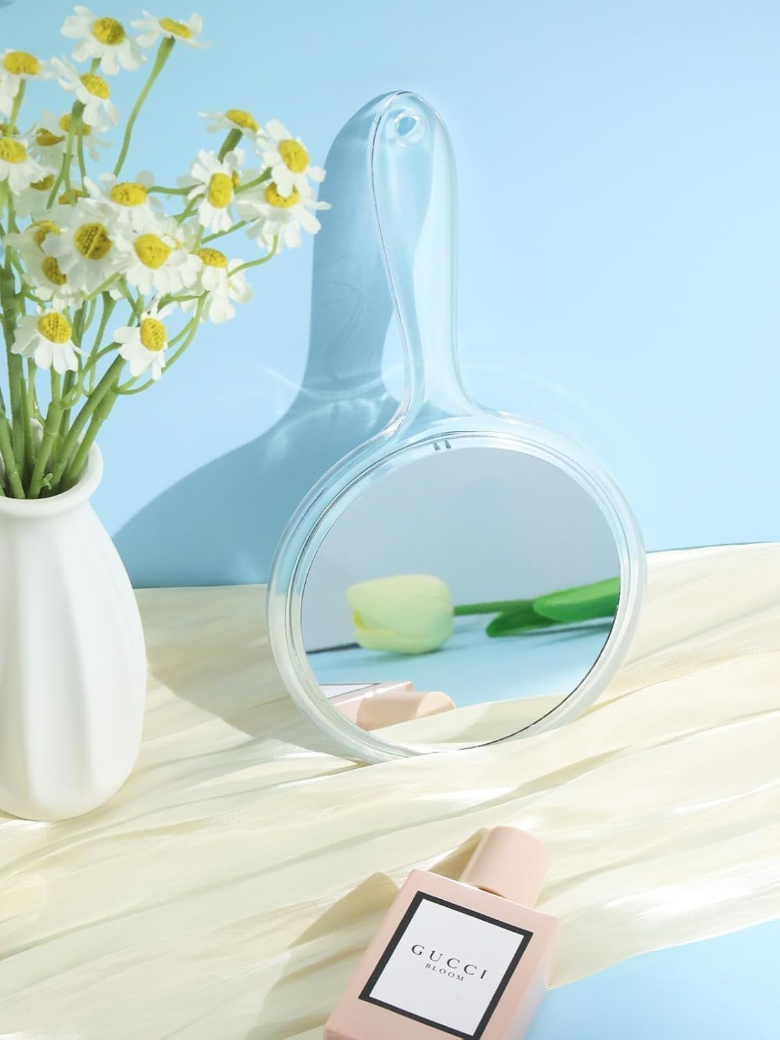 Ein Handspiegel reflektiert eine Tulpe und einen Parf ümflakon. Blumen in einer Vase befinden sich links. Cremefarbener Stoff und hellblauer Hintergrund.