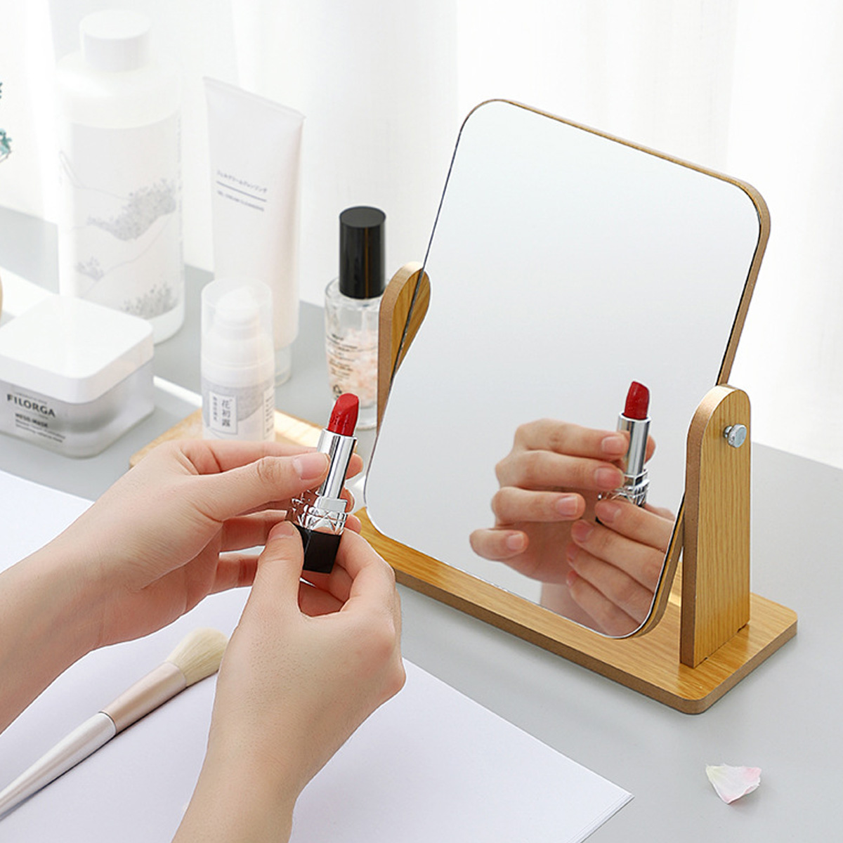 Eine Person hält einen roten Lippenstift vor einen Spiegel. Der Spiegel reflektiert den Lippenstift. Verschiedene Schönheitsprodukte liegen auf dem Tisch.