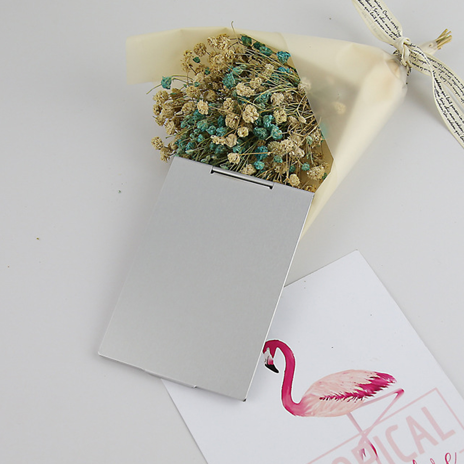 Ein Strauß getrockneter Blumen, eine silberne Karte und eine Karte mit einer Flamingodarstellung auf weißer Oberfläche.