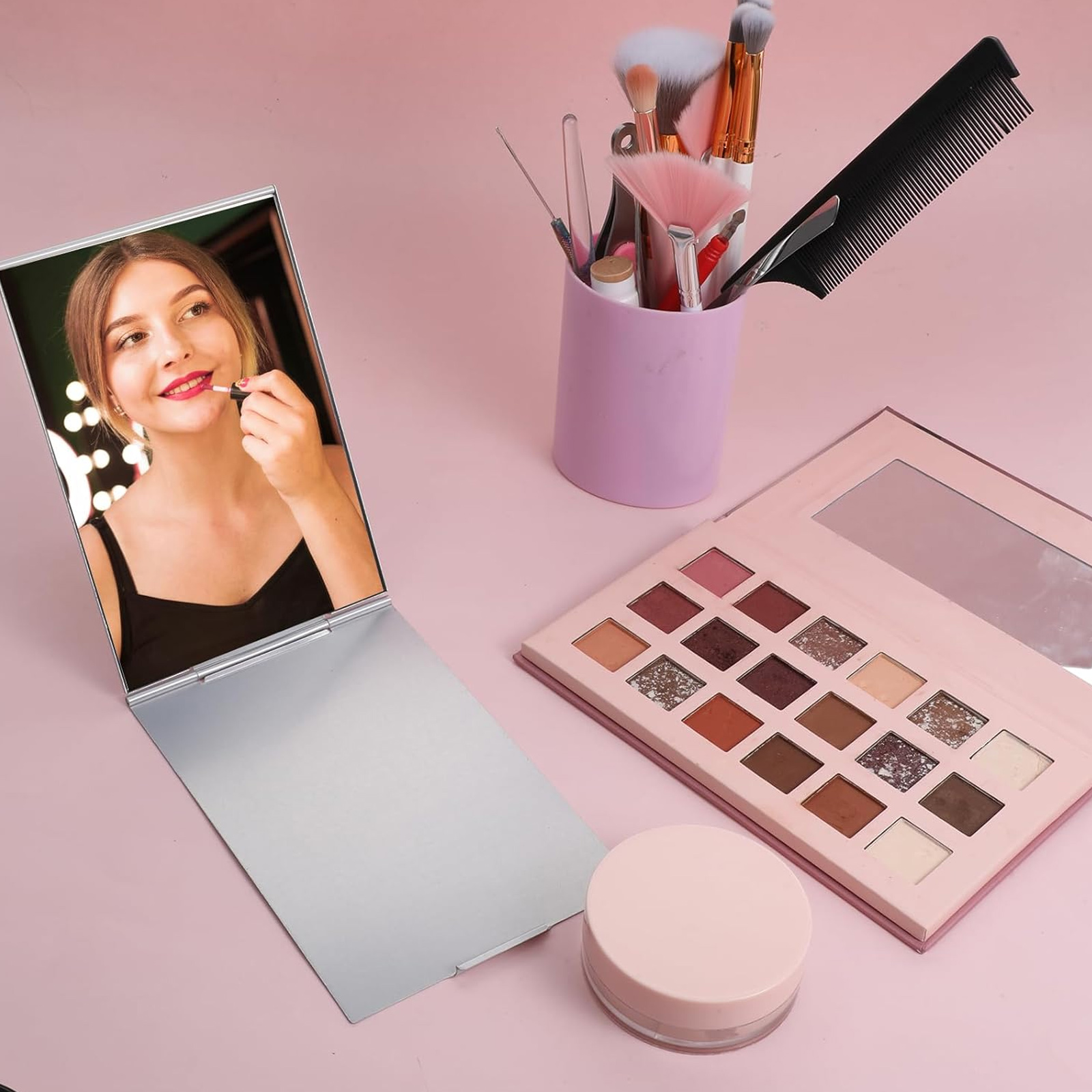 Eine Frau trägt Lippenstift im Spiegel auf. Pinsel, eine Palette und ein Tiegel liegen auf rosa Untergrund.