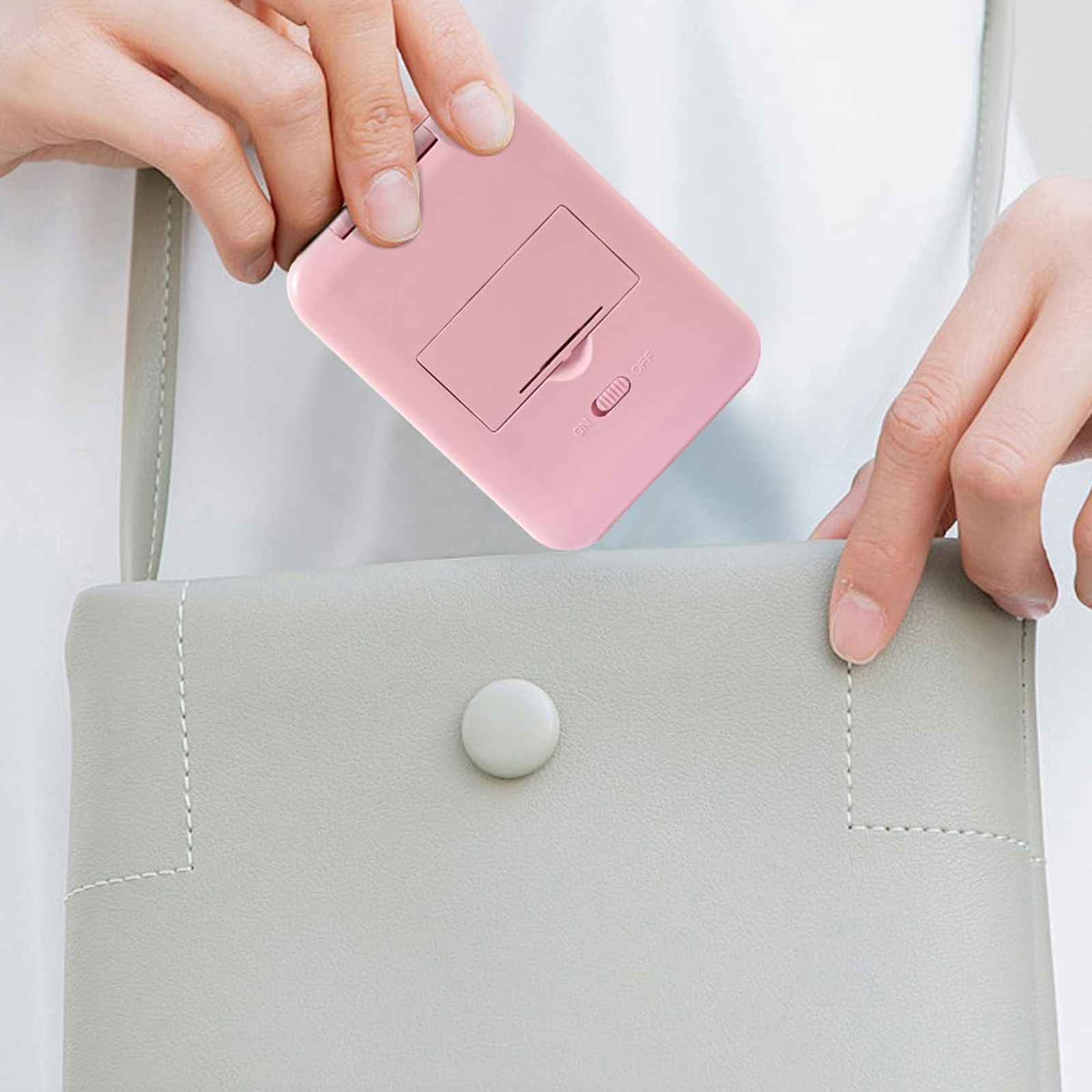 Eine Hand hält ein rosa Gerät mit Ein-/Ausschalter. Es wird in eine graue Handtasche gelegt.